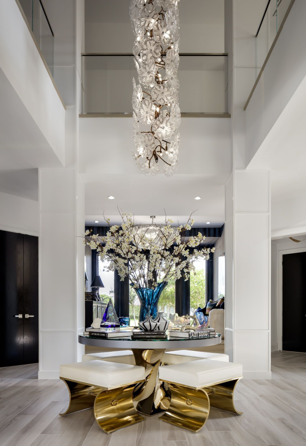 Lori Morris Design - an entryway with a large suspension lamp on the ceiling, a round table with four benches in gold and white details. A view to the back of the house and a living room.