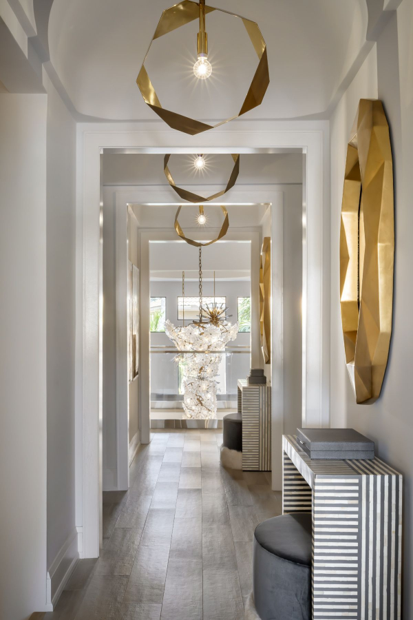 Lori Morris Design - A large hallway with a view on the end to the entryway with two doors on the left, two console and two mirrors on the righ. Three suspension lamps along the hallway. The house has white and grey tones and the furniture is in gold and grey details.
