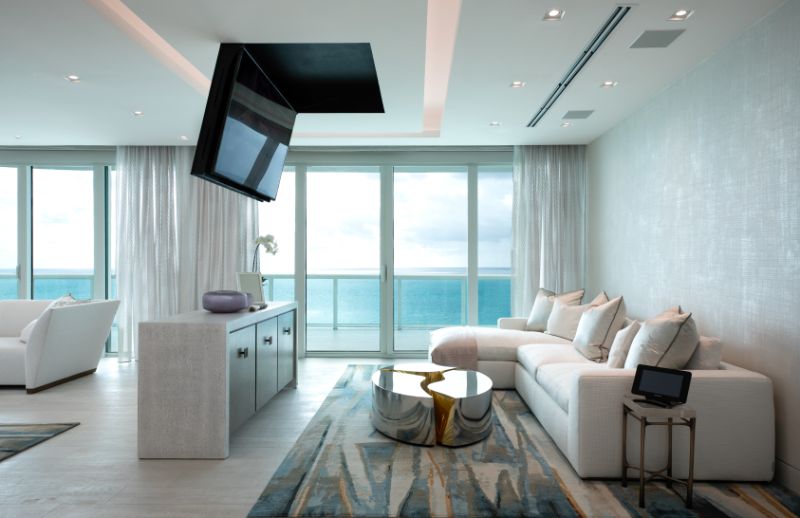 Sarah-Z-Designs - A modern house with a view to the ocean. The house has light tones, a white sofa, a center table in stainless steel, a sideboard and a tv on the ceiling.