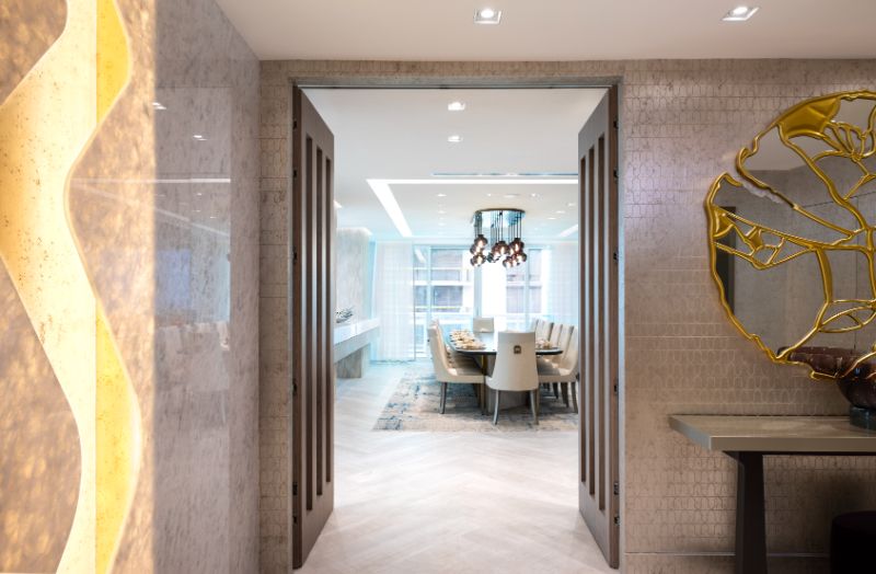Sarah-Z-Designs - Grey tones in the hallway with a console and a mirror with gold details on the wall. A view to the dining space with a rug, a dining table and dining chairs with a chandelier on the ceiling.