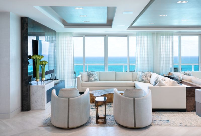 Sarah-Z-Designs - A room that as a view to the ocean, a golden center table, two armchairs in white tones, a white sofa and a large tv