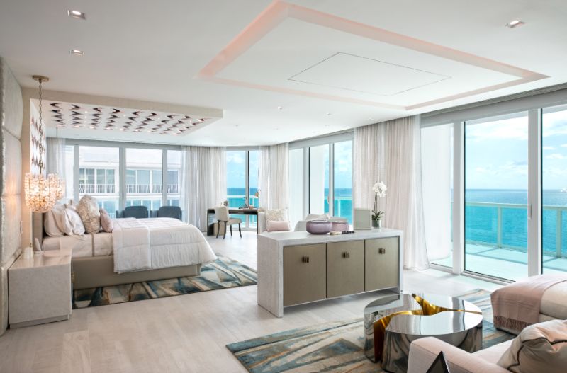 Sarah-Z-Designs - A modern house with a view to the ocean. The house has light tones, a bed, a sofa, a center table in stailess steela and a sideboard.