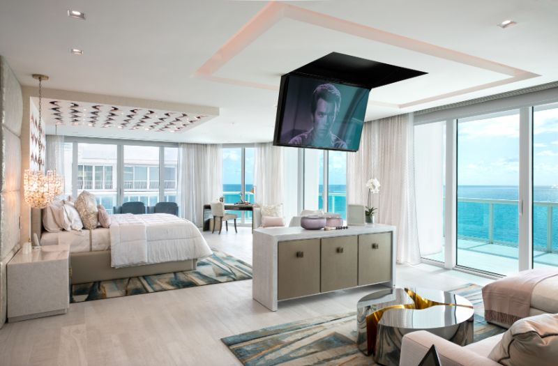 Sarah-Z-Designs - A modern house with a view to the ocean. The house has light tones, a bed, a sofa, a center table, a sideboard and a tv on the ceiling.