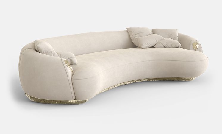 Rockwell Group - A curved sofa in cream fabric. As details in gold on the base and on both arms.